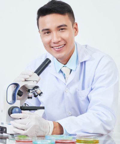 Portrait of happy young Vietnamese chemist looking at various petri dishes through microscope