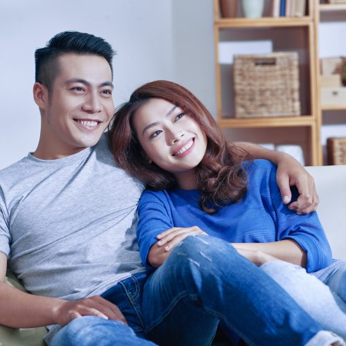 happy-asian-couple-sitting-couch-together-home-looking-away-smiling